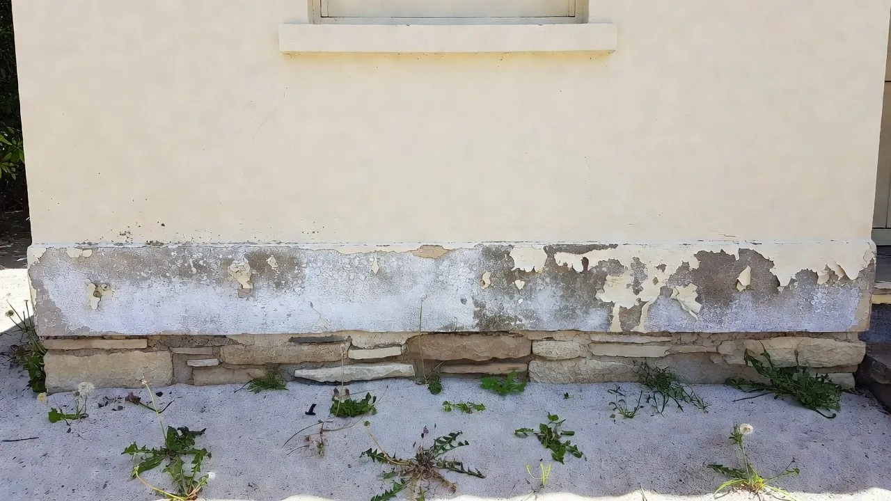 White salt bloom and lifting paint along the bottom of a painted render wall, above a limestone footing.